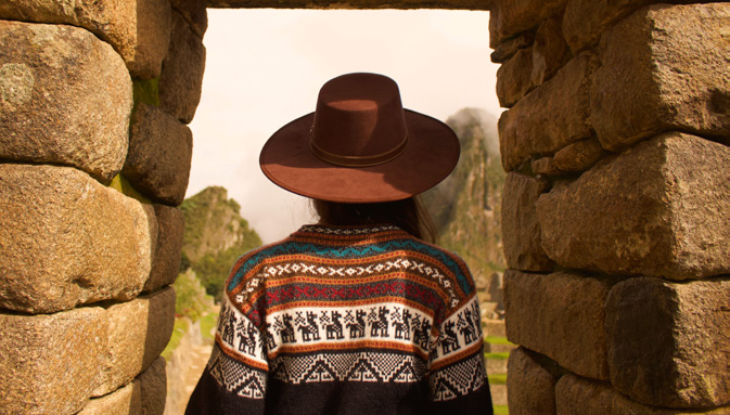 Pessoa de chapéu e poncho tradicional contemplando a paisagem de montanhas através de um portal de pedra